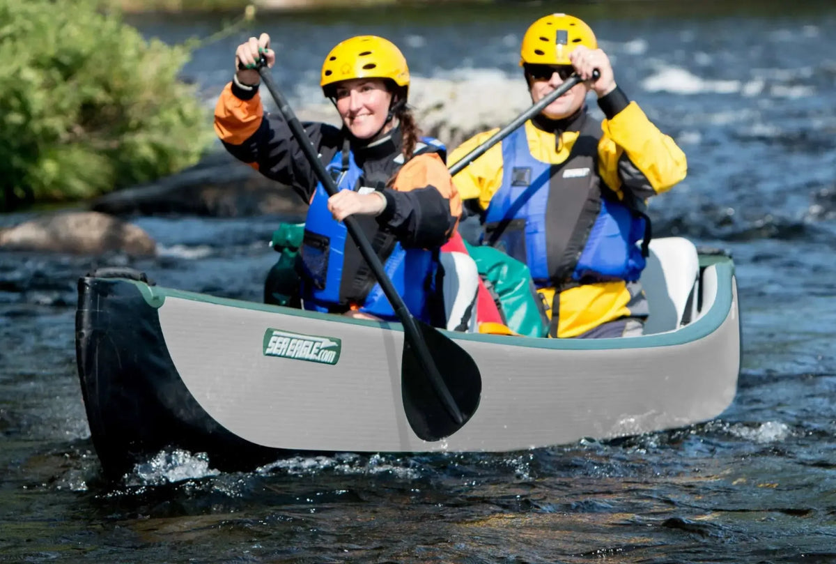 Two paddlers in yellow helmets and life vests in a gray and black Sea Eagle TC16 inflatable canoe on a river.