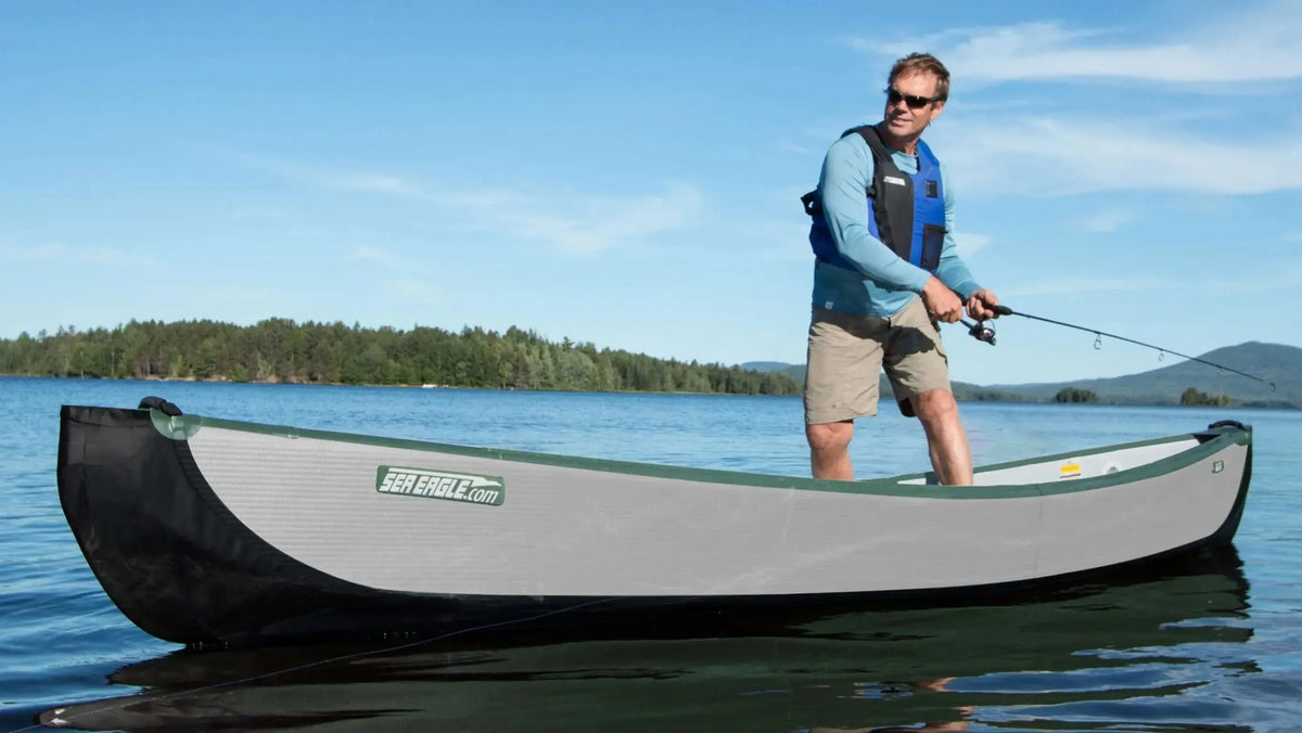 Man standing and fishing from a gray and black Sea Eagle TC16 inflatable canoe on a calm lake.