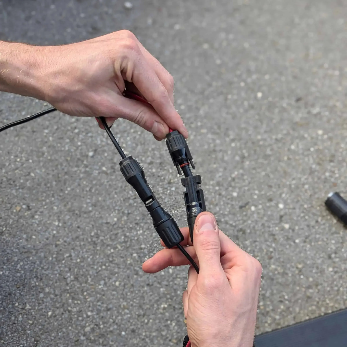 Two hands holding black electrical connectors against a gray pavement background