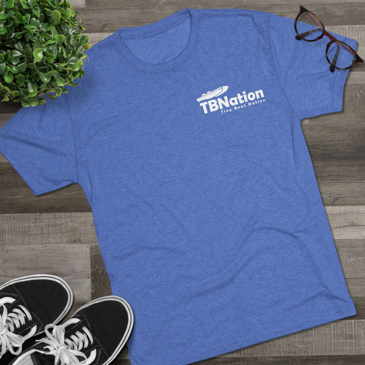 Blue t-shirt with 'TBNation' logo on a wooden surface with sneakers and glasses.