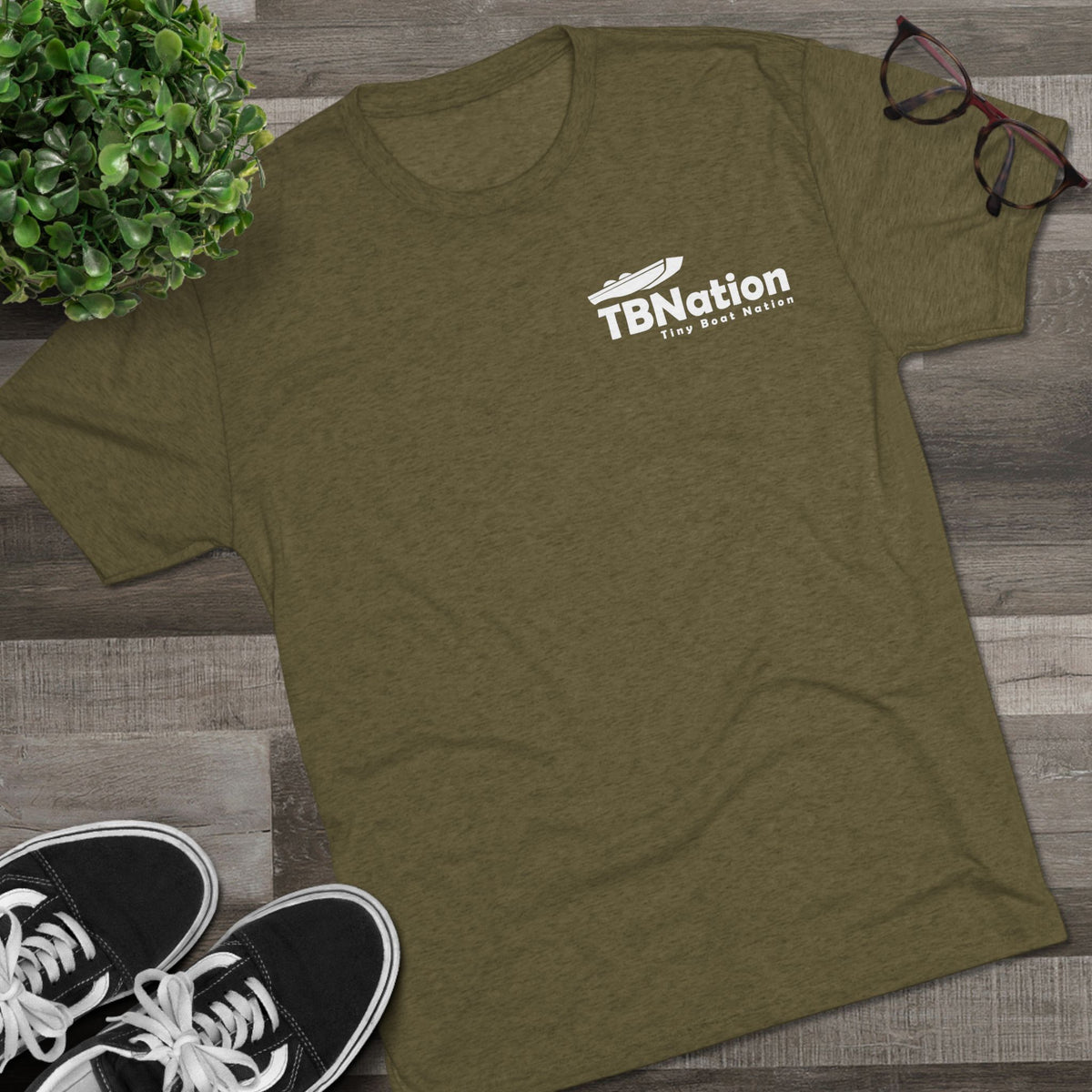 Green t-shirt with a logo on a wooden surface next to sneakers and glasses.