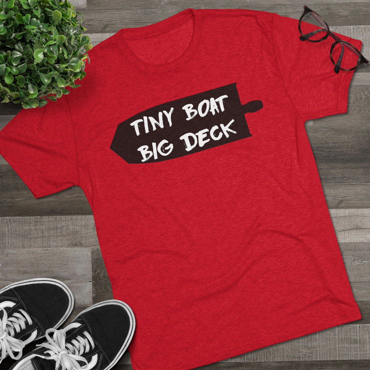 Red t-shirt with 'Tiny Boat Big Deck' text on a wooden surface with shoes and a plant.