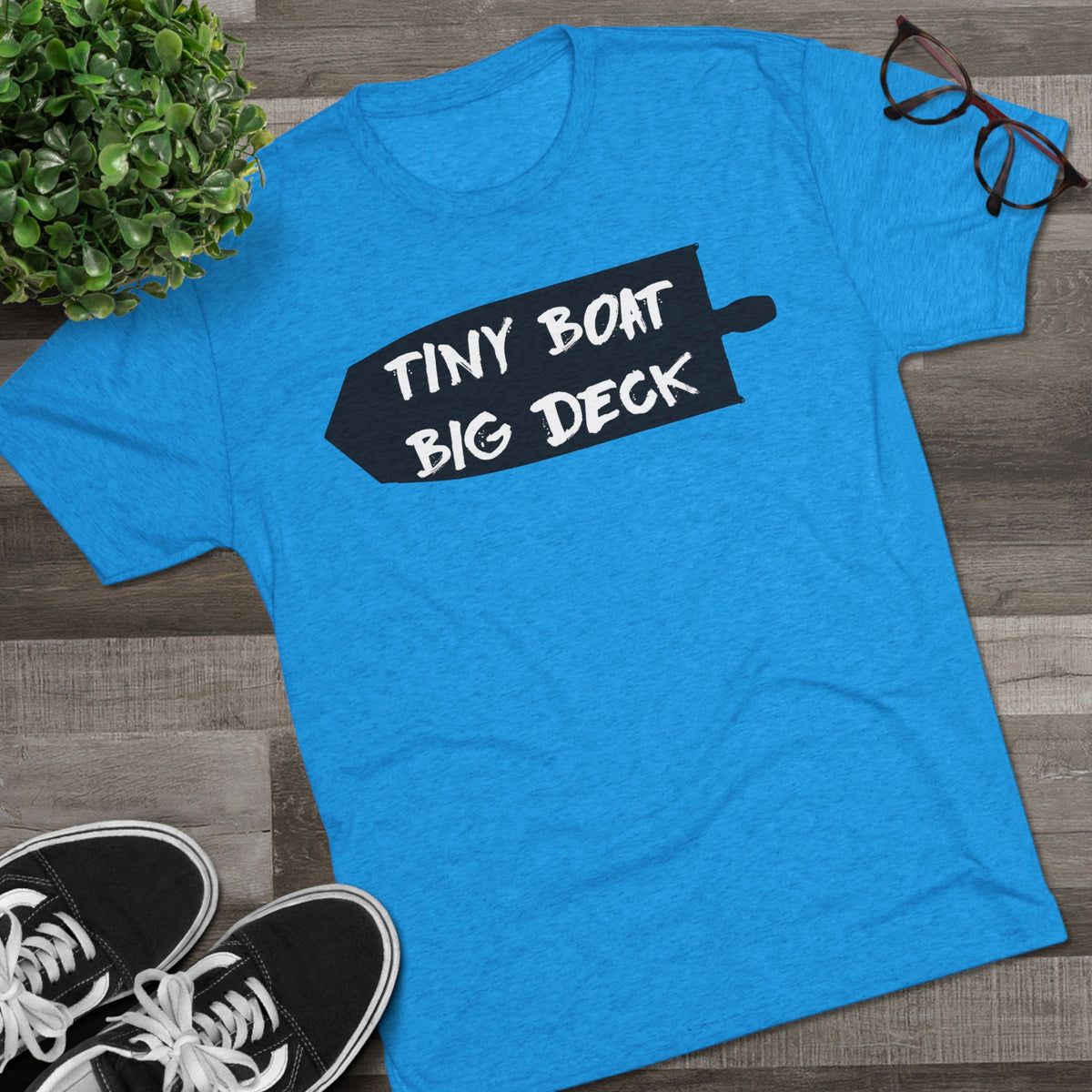 Blue t-shirt with 'Tiny Boat Big Deck' text on a wooden surface with shoes and a plant.