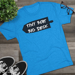 Blue t-shirt with 'Tiny Boat Big Deck' text on a wooden surface with shoes and a plant.