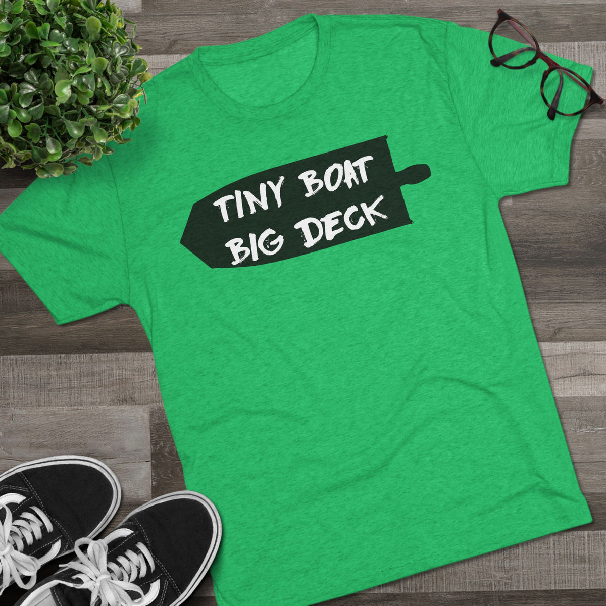 Green t-shirt with 'Tiny Boat Big Deck' text on a wooden surface with shoes and a plant.