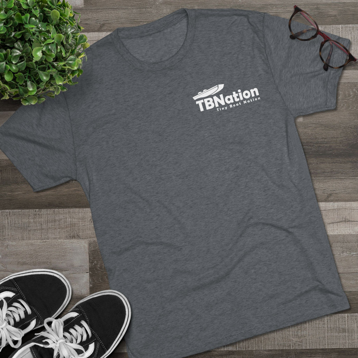 Gray t-shirt with 'TBNation' logo on a wooden surface with sneakers and a plant.