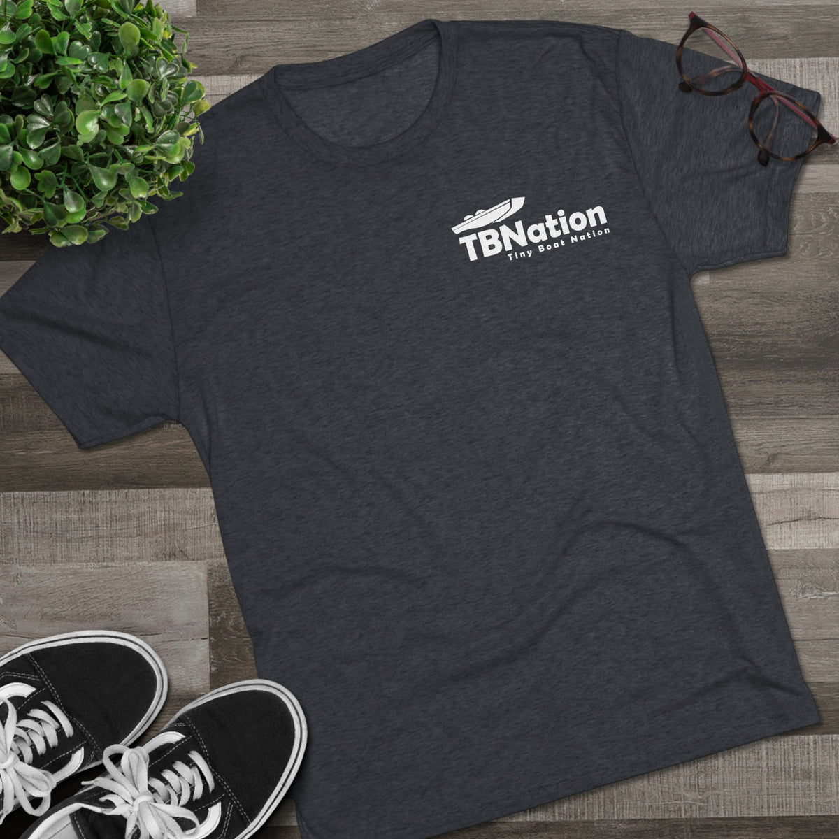 Dark gray t-shirt with 'TBNation' logo on a wooden surface with sneakers and a plant.