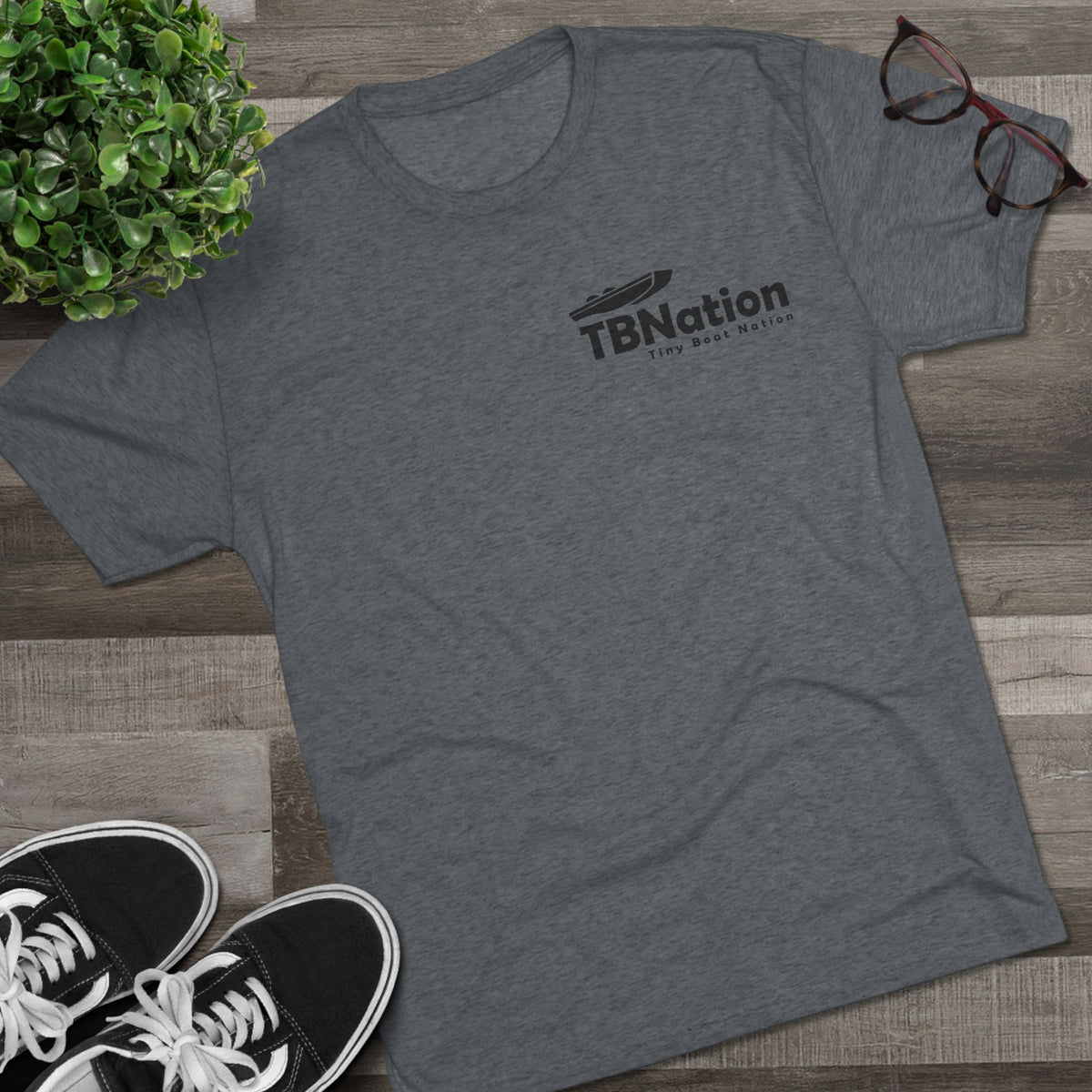 Gray t-shirt with 'TBNation' logo on a wooden surface with sneakers and a plant.