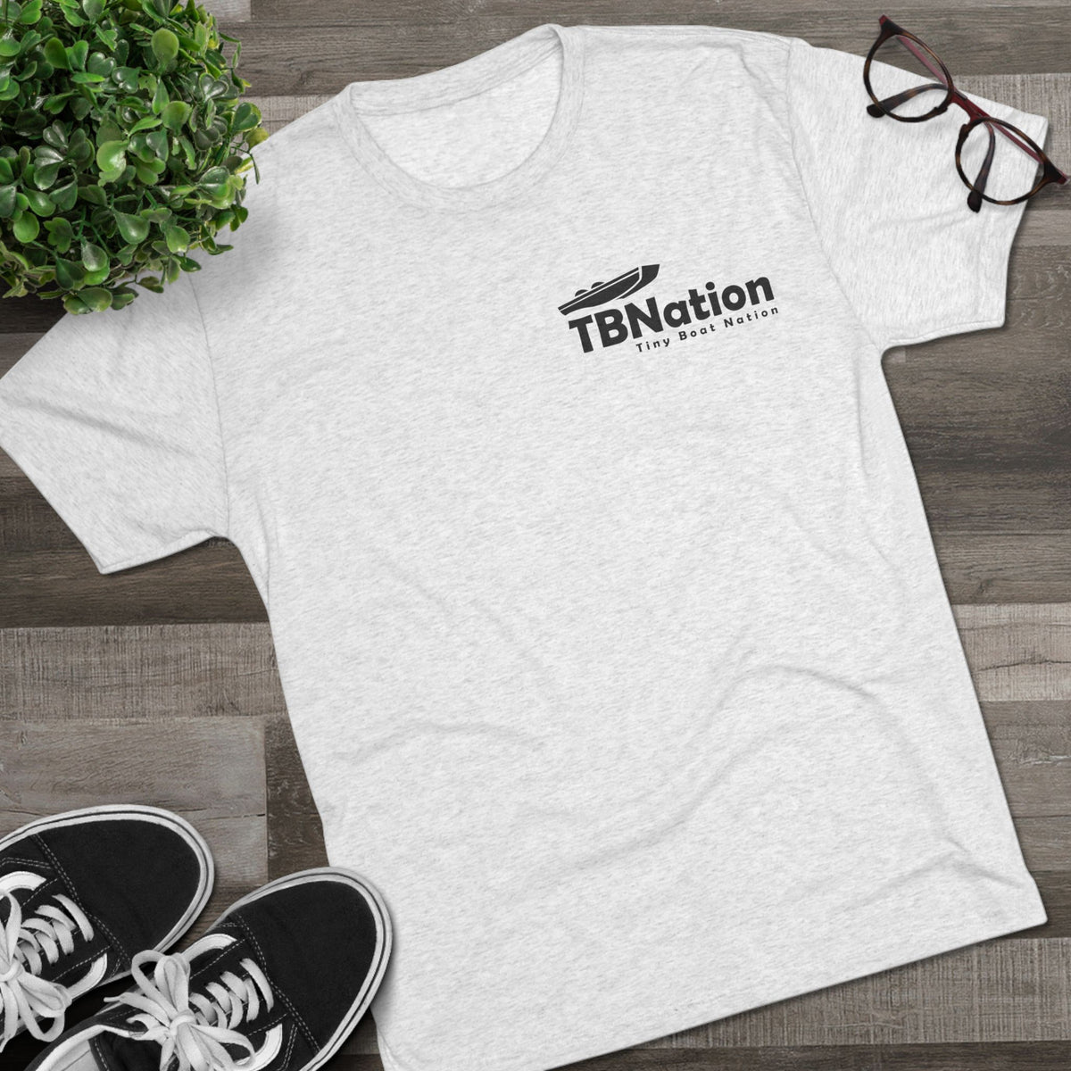 White t-shirt with 'TBNation' logo on a wooden surface with sneakers and glasses.