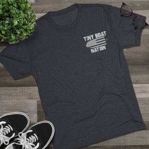 Dark gray t-shirt with 'Tiny Boat Nation' graphic on a wooden surface with sneakers and a plant.