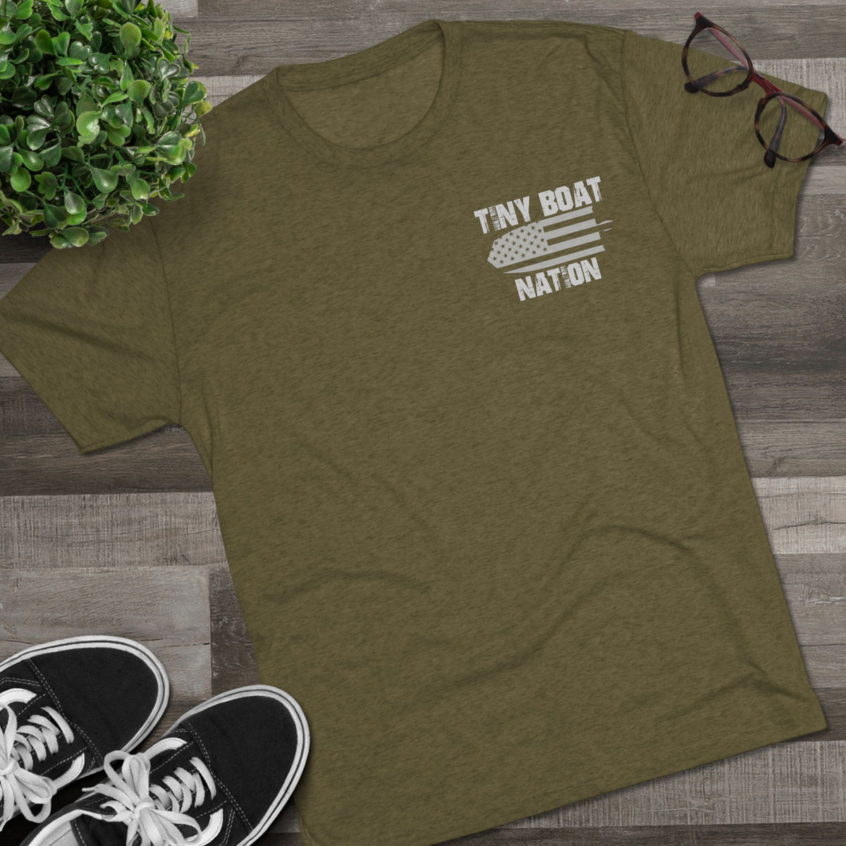 Green t-shirt with 'Tiny Boat Nation' design on a wooden surface with sneakers and a plant.