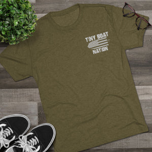 Green t-shirt with 'Tiny Boat Nation' design on a wooden surface with sneakers and a plant.