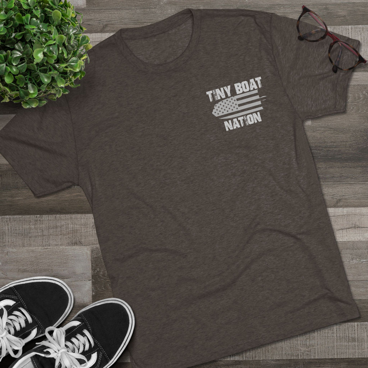 Dark gray t-shirt with 'Tiny Boat Nation' graphic on a wooden surface with sneakers and glasses.