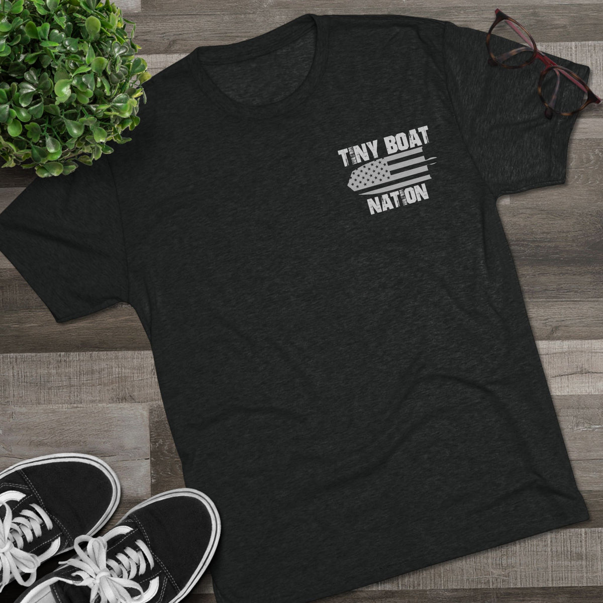 Black t-shirt with 'Tiny Boat Nation' design on a wooden surface with sneakers and a plant.