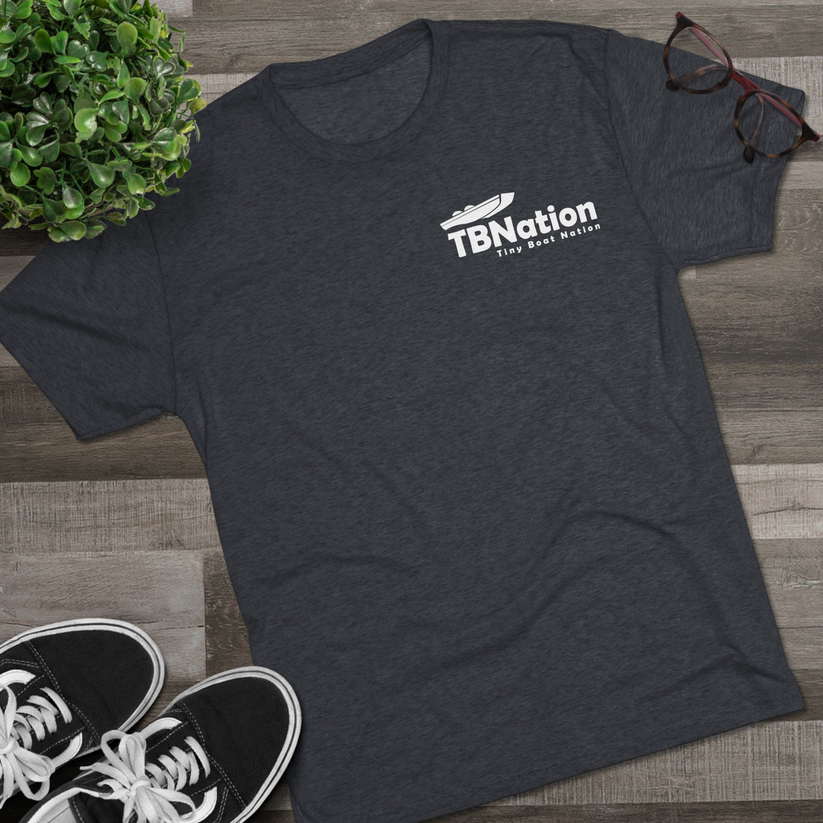 Dark gray t-shirt with 'TBNation' logo on a wooden surface with sneakers and a plant.
