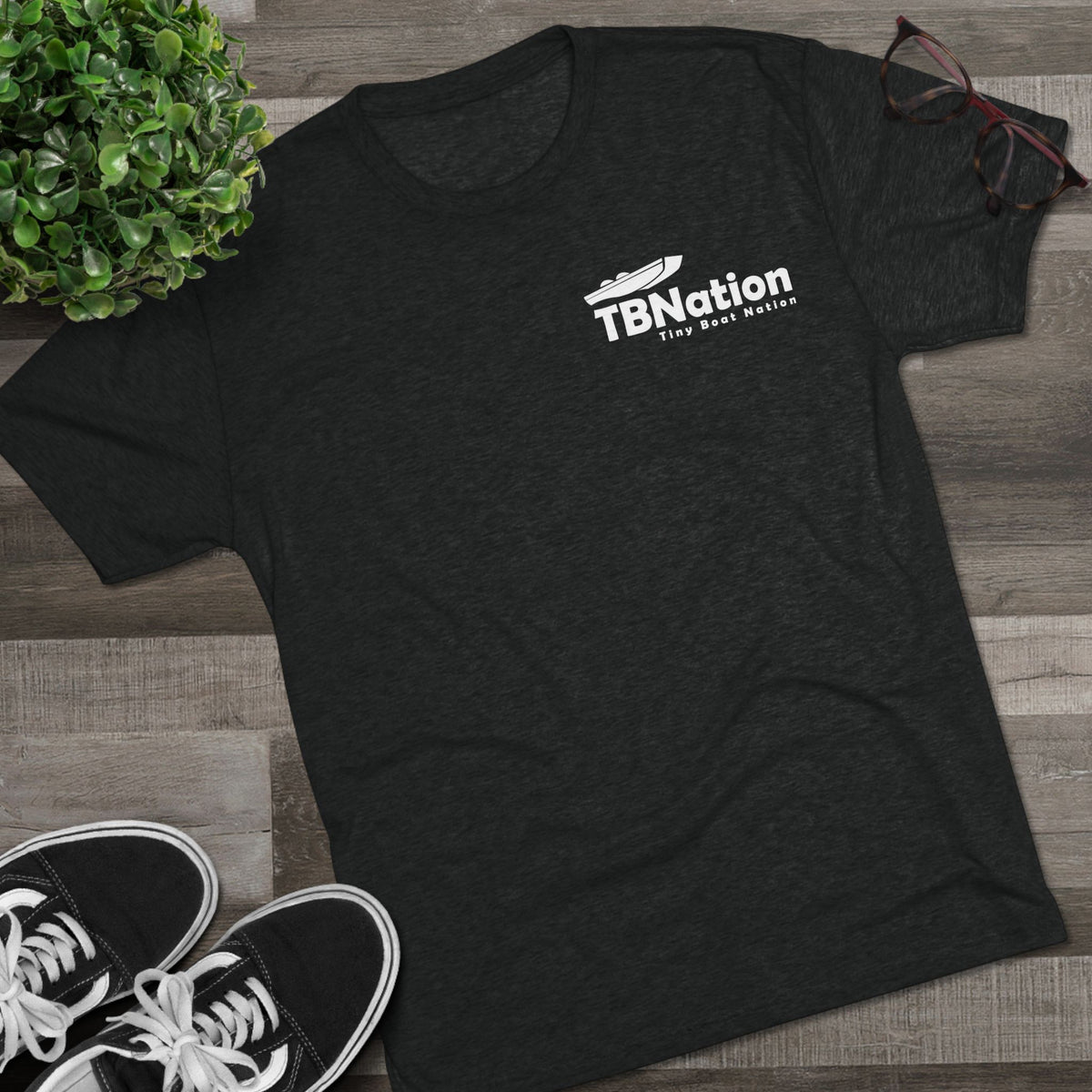 Black t-shirt with 'TBNation' logo on a wooden surface with sneakers and a plant.