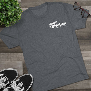 Gray t-shirt with TBNation logo on a wooden surface with sneakers and a plant.