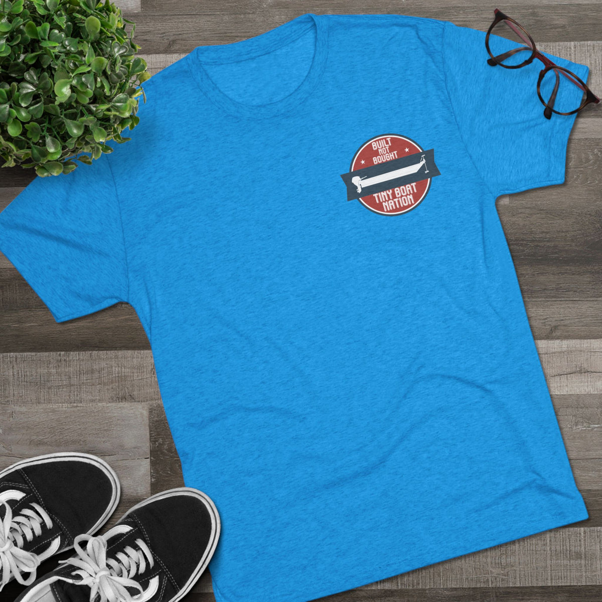 Blue t-shirt with a red and black graphic on a wooden surface with sneakers and a plant.