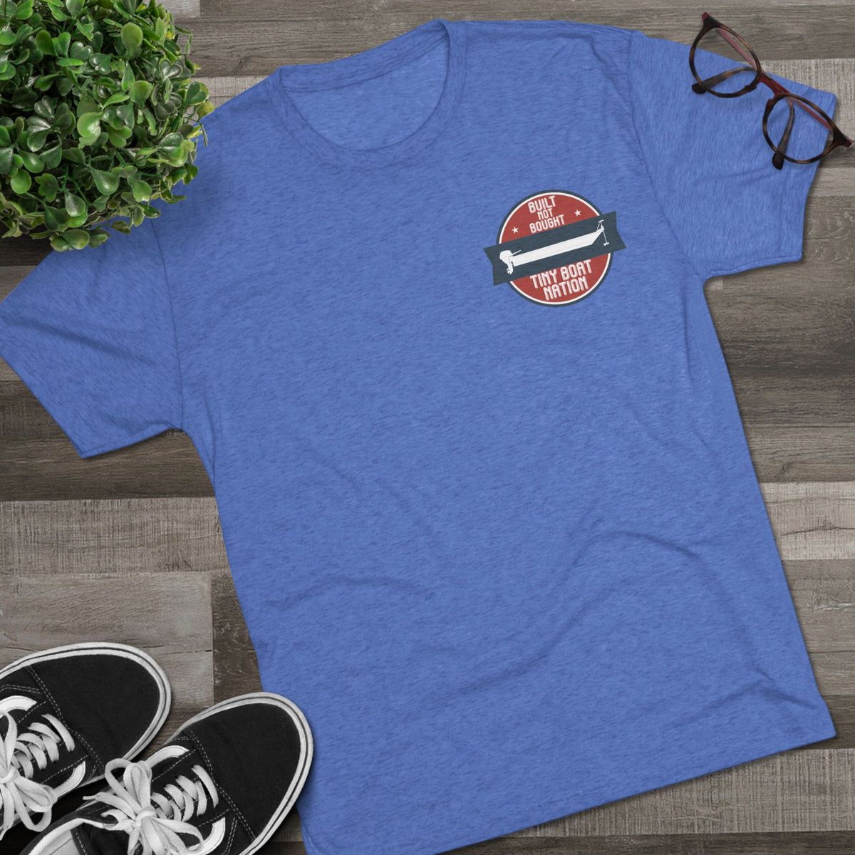 Blue t-shirt with a graphic design on a wooden surface with sneakers and a plant.