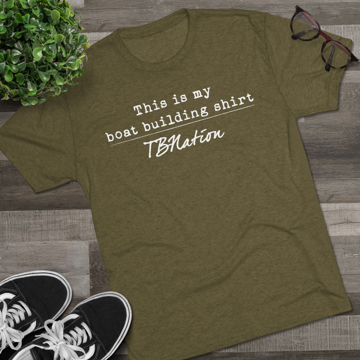 Green t-shirt with text on a wooden surface with shoes and a plant.