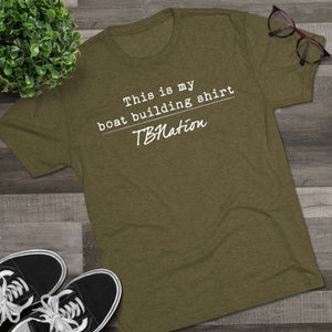 Green t-shirt with text on a wooden surface with shoes and a plant.