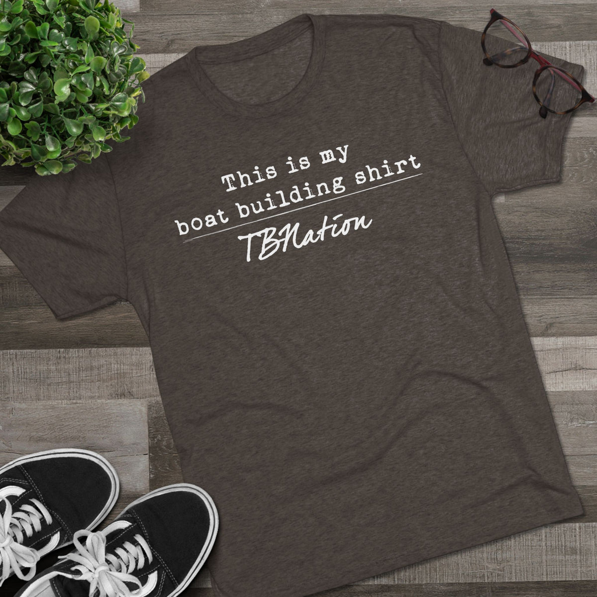 Dark gray t-shirt with text on a wooden surface with sneakers and a plant.