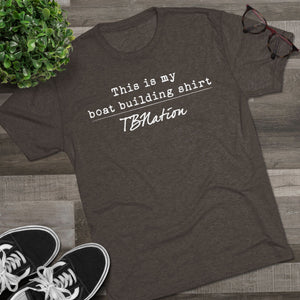 Dark gray t-shirt with text on a wooden surface with sneakers and a plant.