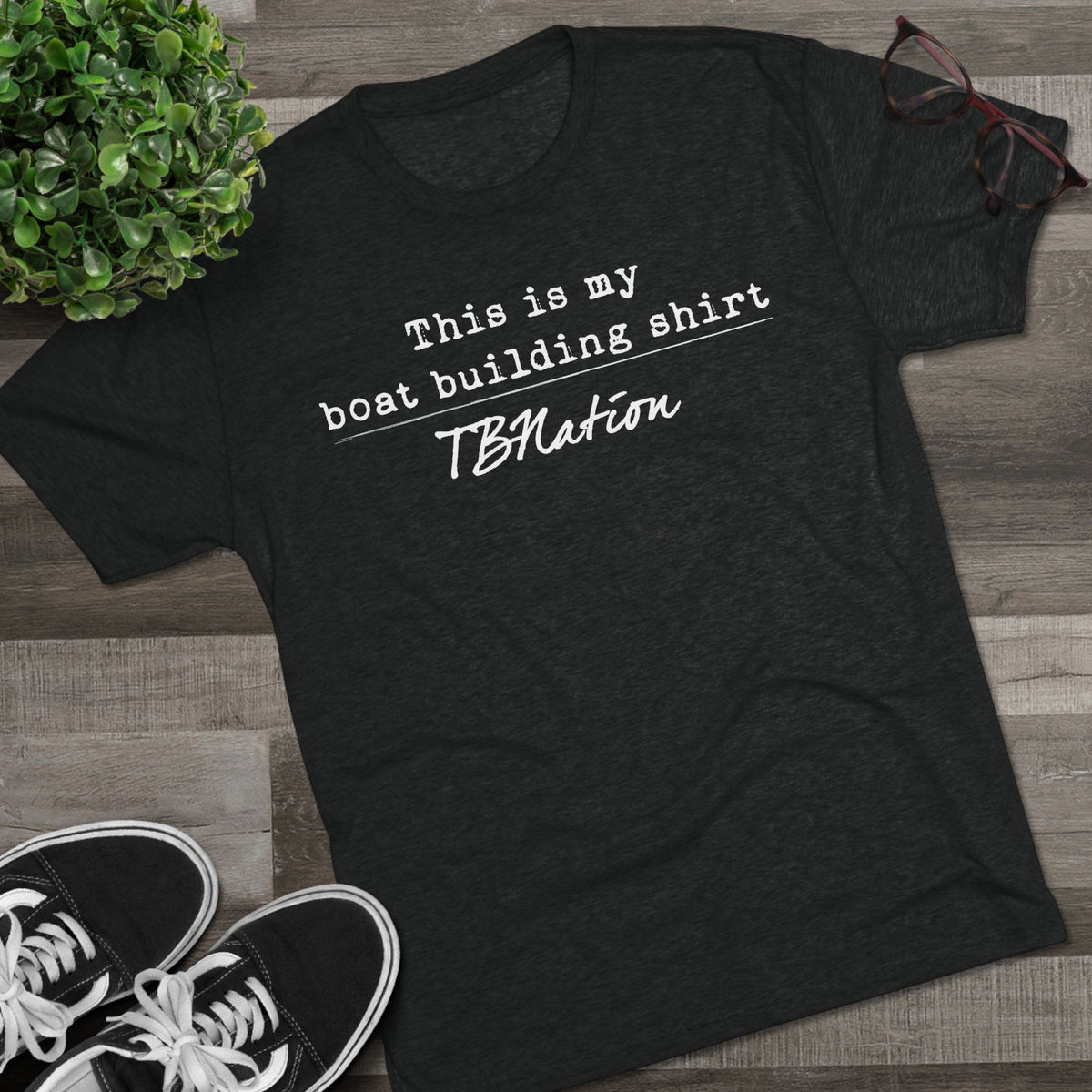 Black t-shirt with text 'This is my boat building shirt TBNation' on a wooden surface with shoes and a plant.