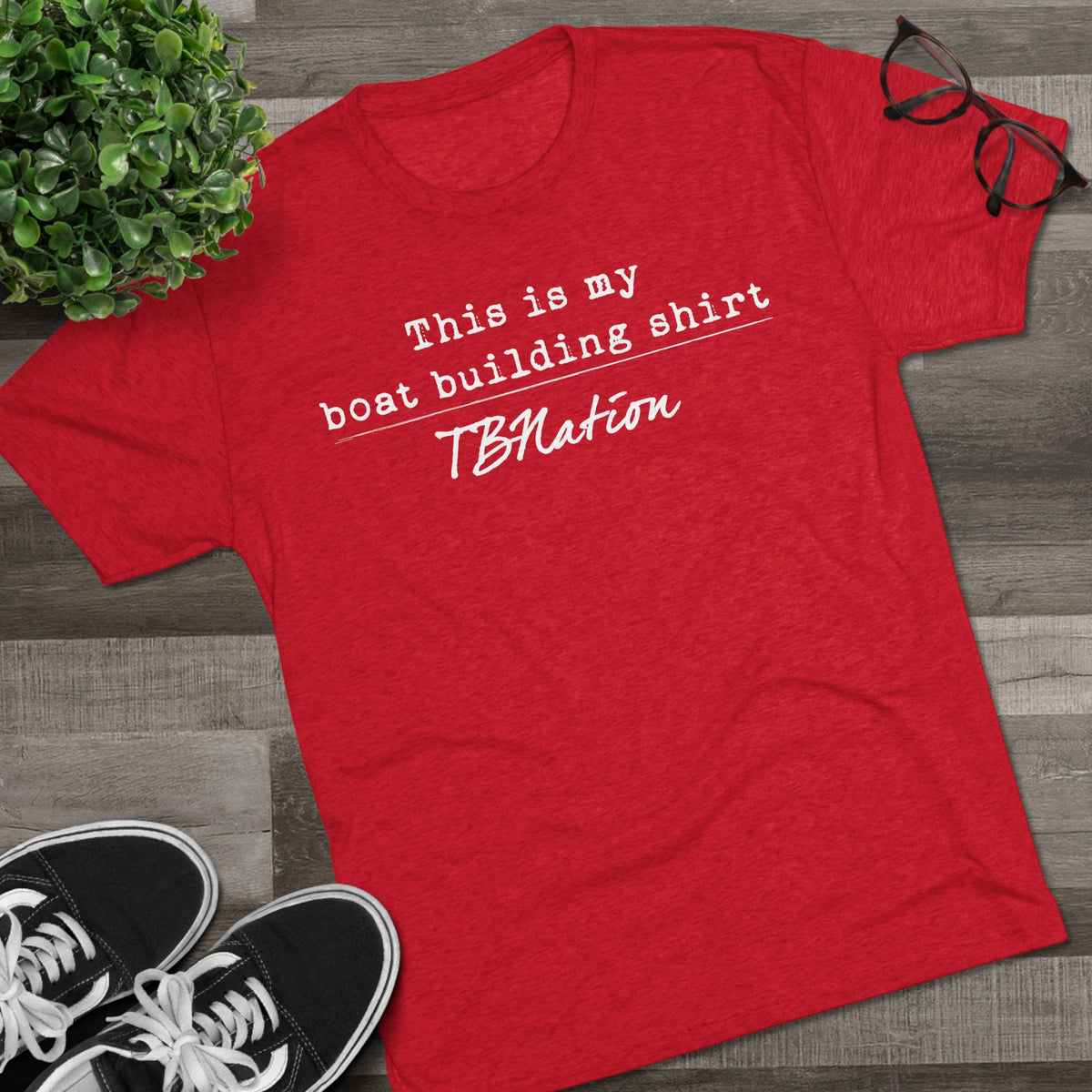 Red t-shirt with text on a wooden surface with shoes and a plant.
