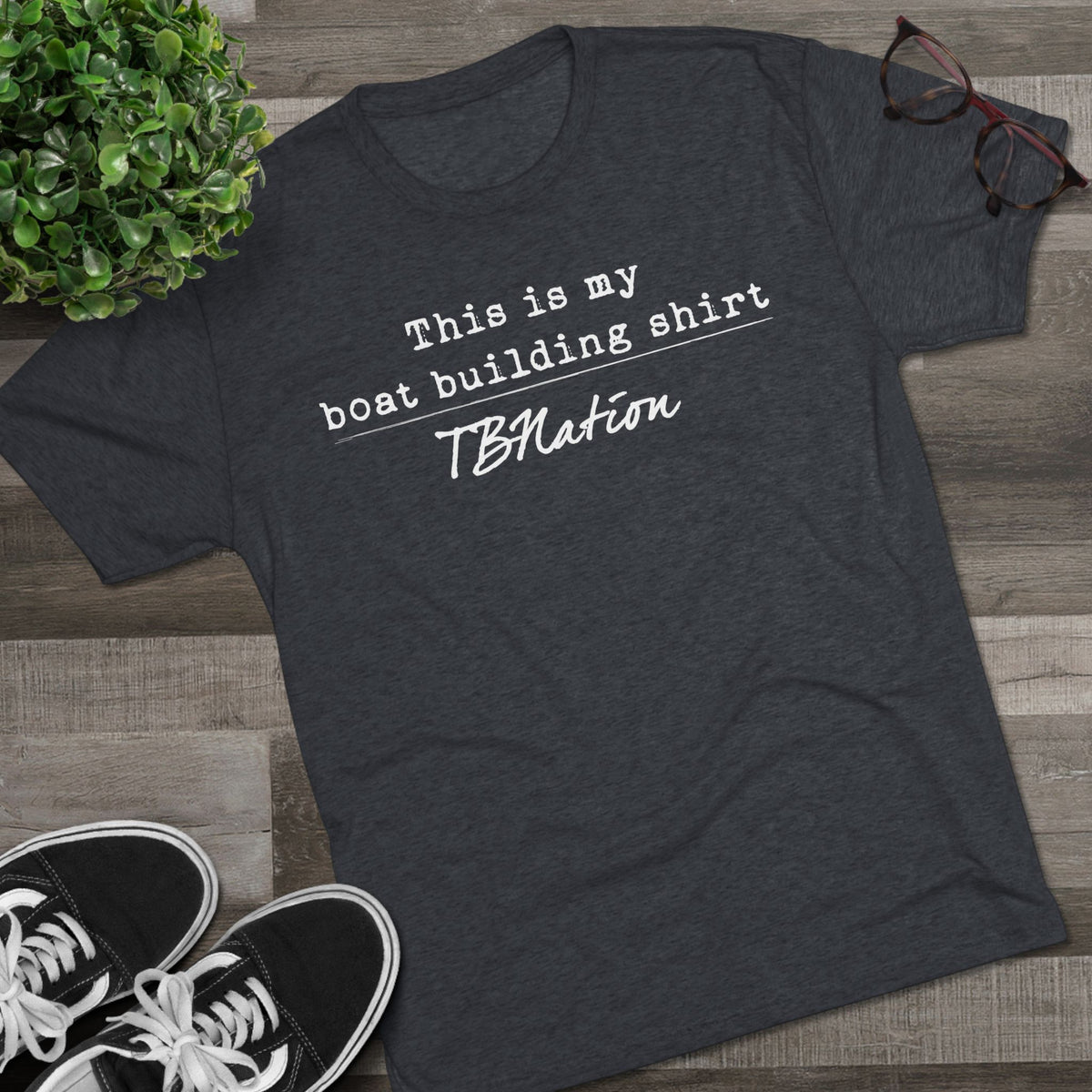 Dark gray t-shirt with text on a wooden surface with sneakers and a plant.