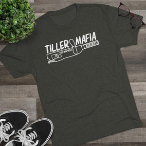 A dark olive green graphic t-shirt laid flat on a gray wood plank surface, styled with a potted green plant, eyeglasses, and black sneakers. The t-shirt features a white graphic that reads 'TILLER MAFIA' with a baseball bat and bottle illustration.