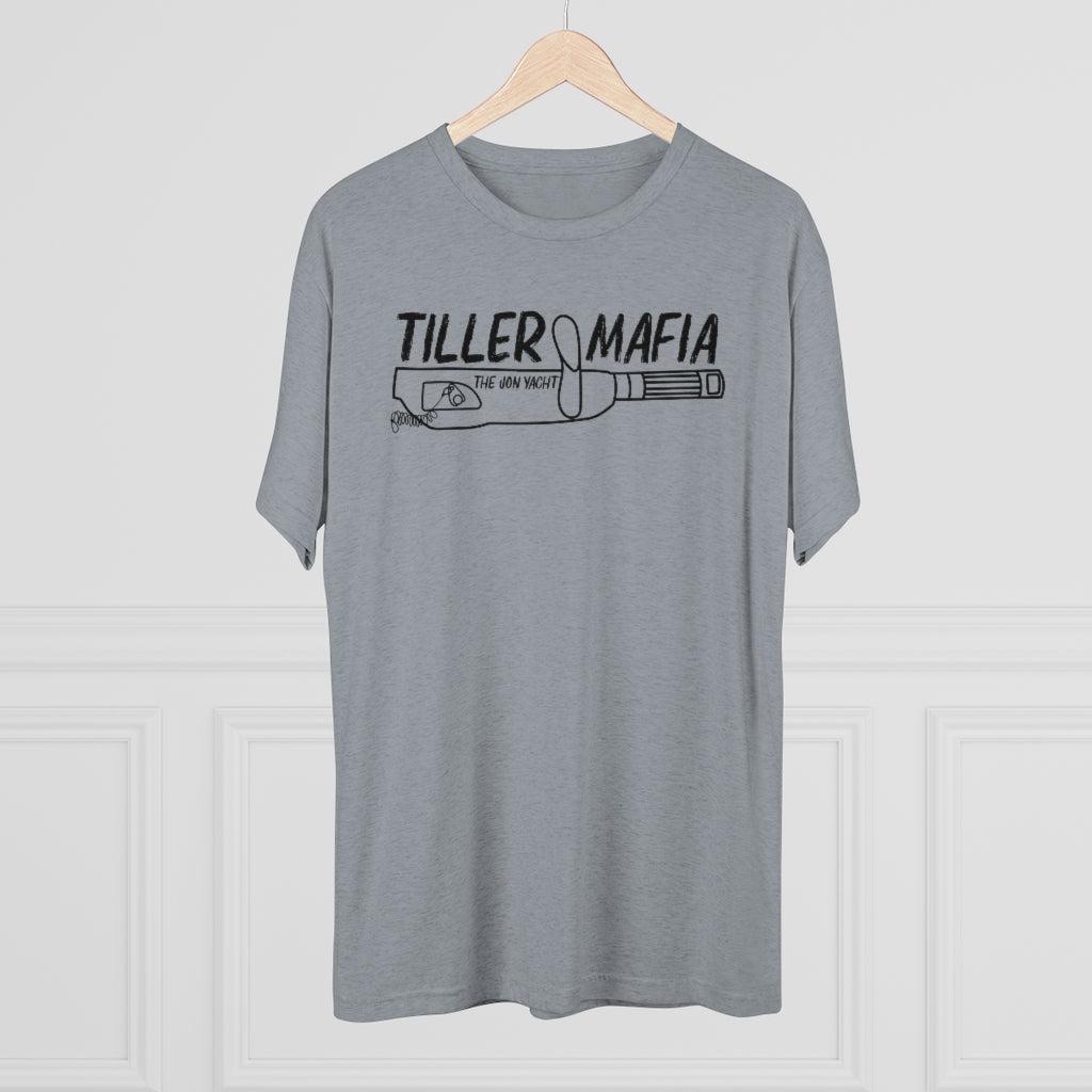 A heather gray short-sleeve t-shirt hanging on a wooden hanger against a white paneled wall. The shirt features a black graphic print on the chest that reads 'TILLER MAFIA' above a drawing of a revolver.