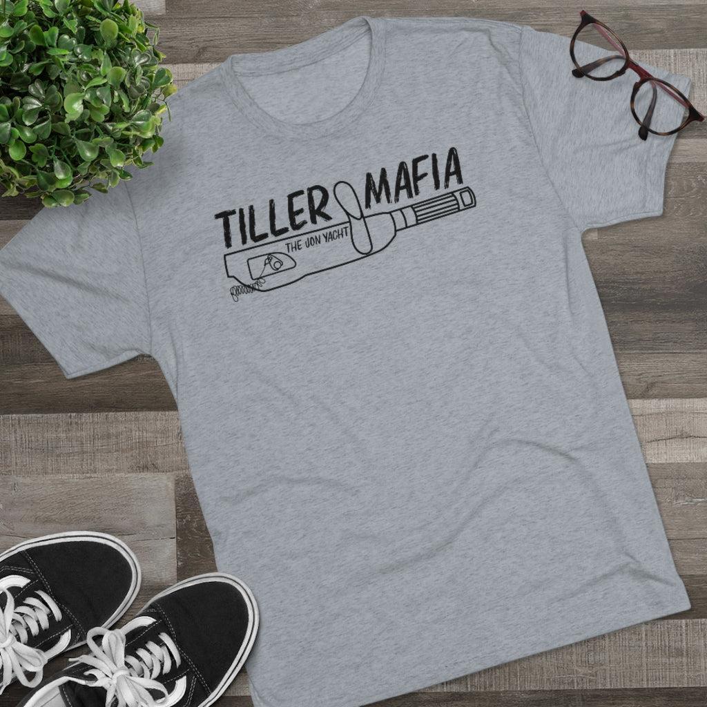 A heather gray short-sleeve t-shirt with a black graphic reading 'TILLER MAFIA' and 'THE JON YACHT' over a baseball bat illustration, styled flat-lay on a wood floor with a plant, glasses, and black sneakers.