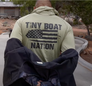 Person wearing a green shirt with 'Tiny Boat Nation' text and American flag graphic outdoors.