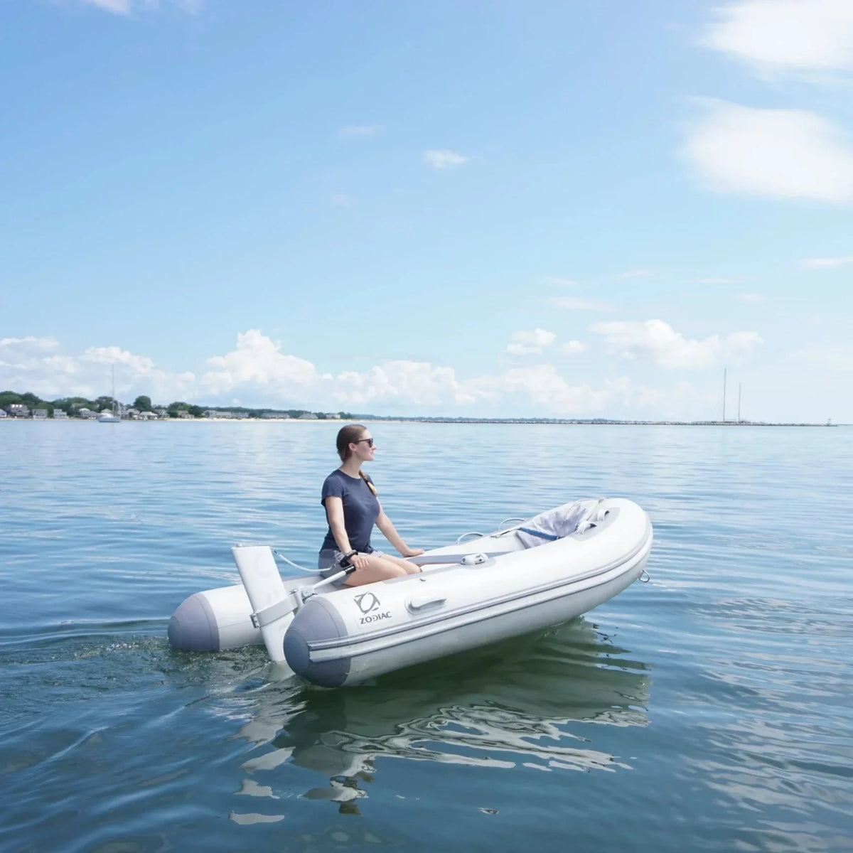 A woman wearing sunglasses and a blue t-shirt sits in a white Zodiac inflatable boat on calm, open water under a bright blue sky. Mounted on the transom is a sleek, white Remigo One electric outboard motor, propelling the boat forward with a gentle wake.