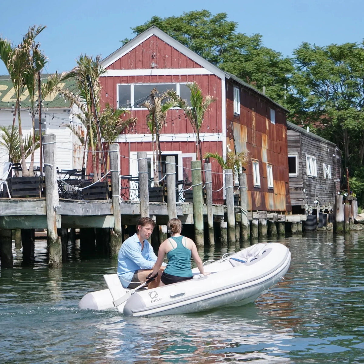 A couple in a Zodiac inflatable boat cruises past a rustic red wooden building on stilts in a harbor. The Remigo One electric outboard motor is visible at the rear, moving the boat quietly through the water near the dock.