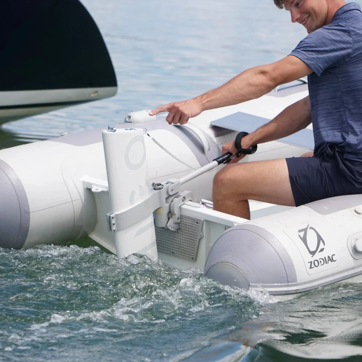 A close-up action shot of a person operating the Remigo One electric outboard. One hand holds the long tiller handle for steering, while the other points toward the motor’s sleek, integrated housing. The boat is creating a visible splash as it moves through the water.