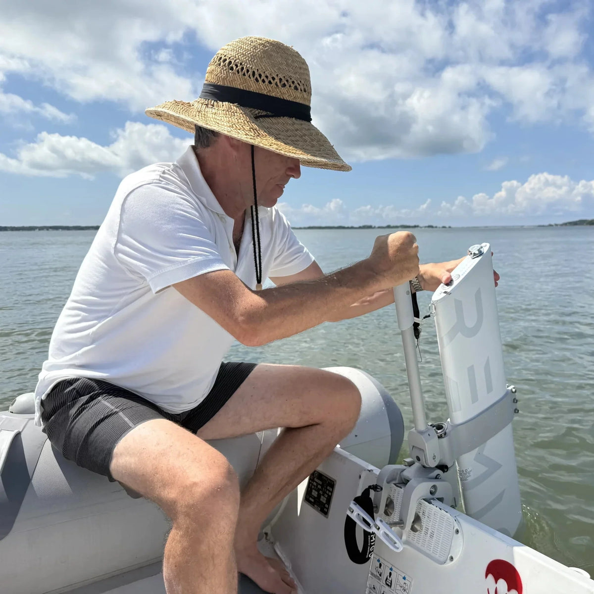 A man wearing a straw hat and white polo shirt sits in an inflatable boat, reaching back to adjust or secure the mounting bracket of a Remigo One electric outboard. The motor's vertical, pillar-like design is clearly visible against the water.