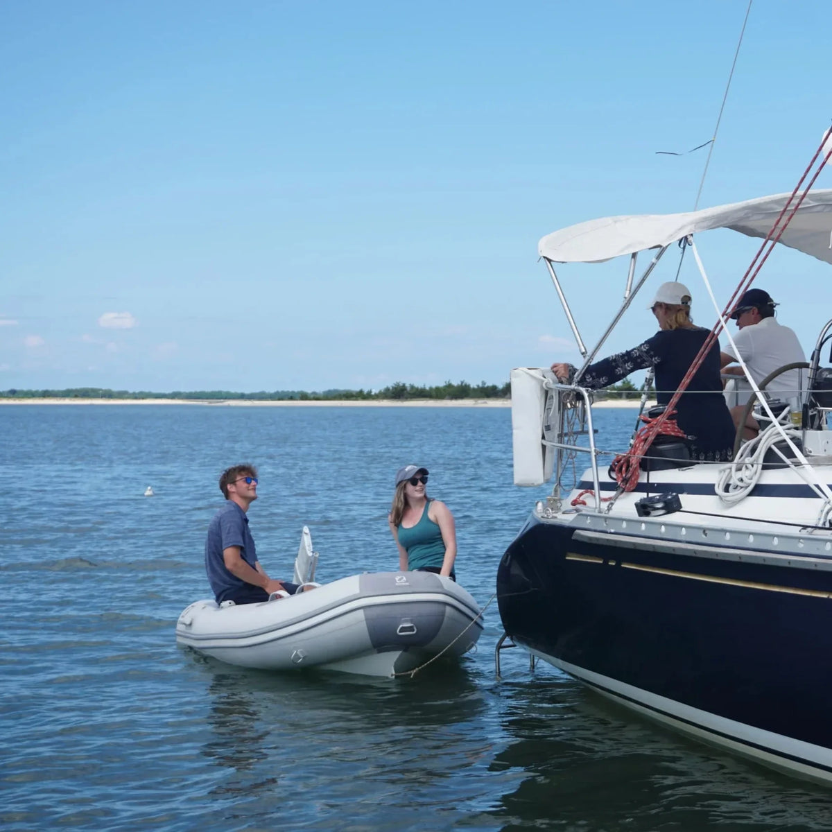 Two people in a small white inflatable tender powered by a Remigo electric outboard pull alongside a large sailboat. The scene is set in a calm harbor with a distant shoreline under a clear sky, highlighting the motor's use as a sailboat tender.
