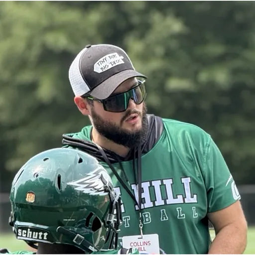 Kyle Henschel, TBNation team member, wearing sunglasses and a cap while standing beside football equipment, representing leadership, teamwork, and a passion for the outdoors.
