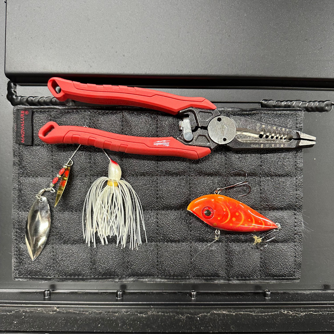 Black quilted Magnalure Pad holding red pliers, a silver spinnerbait, a white jig, and an orange crankbait.