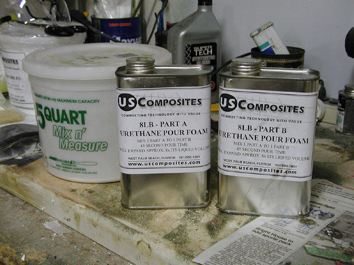 Two silver US Composites 8lb urethane pour foam cans (Part A &amp; B) next to a white 5-quart measuring bucket.